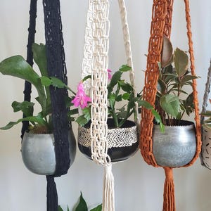 May include: A collection of macrame plant hangers in black, cream, and rust-orange. Each hanger holds a metal or ceramic pot with a plant. The plants have green leaves and some have pink flowers. The hangers are suspended against a white wall.