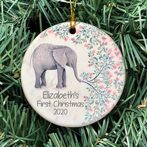 May include: A white ceramic Christmas ornament with a gray elephant illustration and pink and green floral design. The ornament reads "Elizabeth's First Christmas 2020".