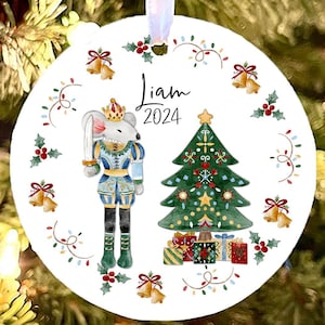May include: White round Christmas ornament with a watercolor illustration of a nutcracker, a Christmas tree, and the text "Liam 2024".