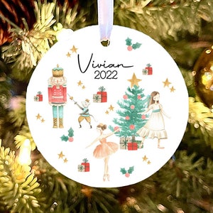 May include: White round Christmas ornament with a watercolor illustration of a Nutcracker, a ballerina, a Christmas tree, and presents. The ornament has the name "Vivian" and the year "2022" written on it.