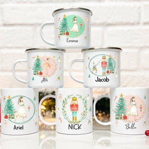 Puede incluir: Seis tazas de esmalte blanco con nombres personalizados, con un tema navideño con una bailarina, un cascanueces y un árbol de Navidad. Las tazas están dispuestas en semicírculo sobre una superficie blanca.