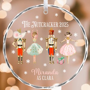 May include: A clear, round Christmas ornament with a white ribbon. The ornament features a Nutcracker theme with illustrations of characters and the text "The Nutcracker 2025" and "Miranda AS CLARA". The characters include a mouse, a ballerina, a nutcracker, and another ballerina.