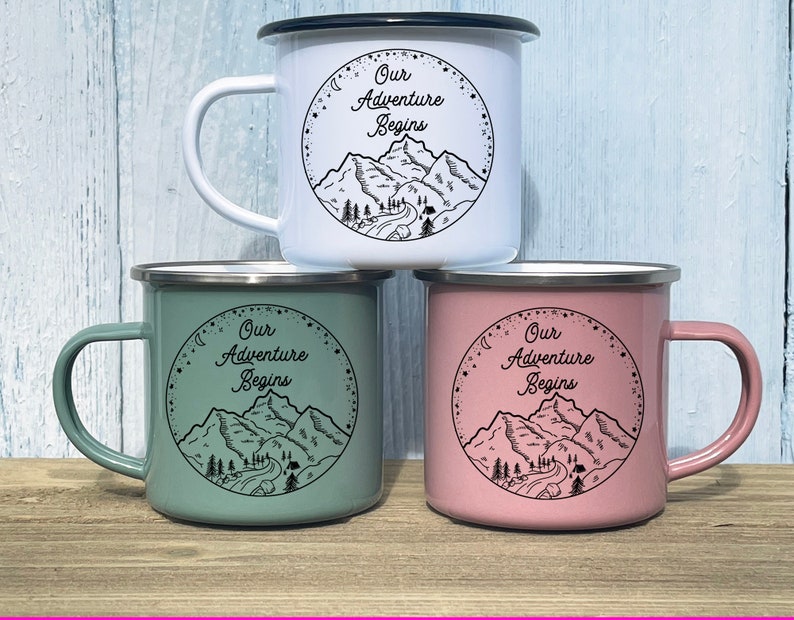 K&ouml;nnte beinhalten: Drei Emailletassen mit schwarzen R&auml;ndern und Griffen. Die Tassen sind wei&szlig;, gr&uuml;n und rosa. Jede Tasse hat ein schwarz-wei&szlig;es Motiv von Bergen und den Text "Our Adventure Begins".