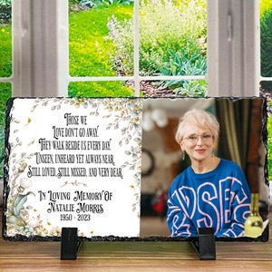 May include: A black slate memorial plaque with a floral design and a photo of a woman. The text reads "Those we love don't go away. They walk beside us every day. Unseen, unheard yet always near. Still loved, still missed, and very dear. In loving memory of Natalie Morris 1950-2023."