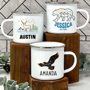 K&ouml;nnte beinhalten: Drei wei&szlig;e Emaille-Campingbecher mit personalisierten Namen und Naturmotiven. Die Becher zeigen ein Reh, einen B&auml;ren und einen Adler mit Bergen und Sternen im Hintergrund. Die Becher stehen auf Holzbl&ouml;cken.