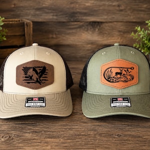 May include: Two adjustable trucker hats with black mesh backs. The left hat is beige with a brown leather patch featuring an eagle and American flag. The right hat is olive green with a tan leather patch showing a deer, duck, and fishing hook.