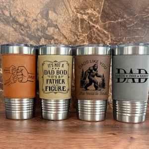 May include: Four stainless steel insulated tumblers with leather sleeves. The sleeves feature various designs and text, including "DAD", "It's Not a Dad Bod It's a Father Figure", and "Dads Like You Are Hard to Find".