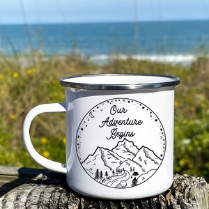 K&ouml;nnte beinhalten: Wei&szlig;e Emaille-Campingtasse mit einer Schwarzwei&szlig;illustration von Bergen, einer Stra&szlig;e und B&auml;umen. Der Text "Our Adventure Begins" ist in einer kursivschriftlichen Schriftart geschrieben.