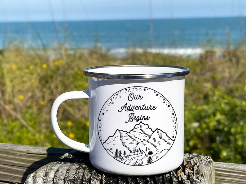 K&ouml;nnte beinhalten: Wei&szlig;e Emaille-Campingtasse mit einer Schwarzwei&szlig;illustration von Bergen, einer Stra&szlig;e und B&auml;umen. Der Text "Our Adventure Begins" ist in einer kursivschriftlichen Schriftart geschrieben.