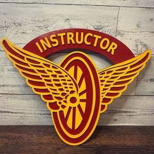 May include: Red and yellow wooden sign with the word "INSTRUCTOR" in a semi-circle above a stylized red and yellow wheel with wings.