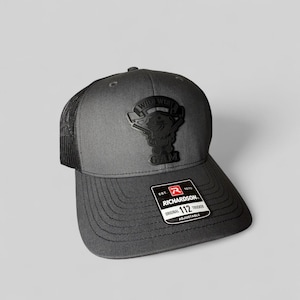 May include: Gray trucker hat with a black mesh back and a curved brim. The hat features a black patch with the words "Wild West" and a cartoon character. The Richardson brand label is visible.
