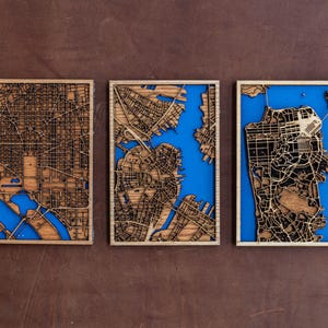 May include: Three wooden wall art pieces featuring laser-cut maps of cities. Each map is framed in a natural wood frame and has a blue water feature.