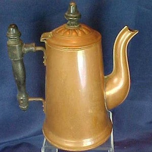 May include: A copper teapot with a wooden handle and a black knob on the lid. The teapot has a shiny, polished finish.