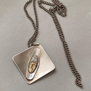 May include: A silver-toned necklace featuring a square pendant with an oval-shaped inlay of iridescent abalone shell. The pendant hangs from a matching chain. The necklace has a vintage aesthetic.