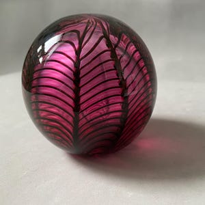 Può includere: Un fermacarte sferico in vetro trasparente con una vibrante tonalità magenta. Linee nere e curve creano un motivo a forma di piuma sulla superficie. Il fermacarte è appoggiato su una superficie bianca, proiettando un'ombra.