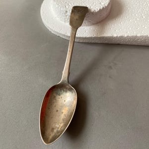 Könnte beinhalten: Ein antiker Silberlöffel mit langem Stiel und ovaler Schale. Der Löffel weist eine leicht angelaufene Oberfläche auf, was auf sein Alter hindeutet. Der Griff hat eine rechteckige Oberseite mit einer Inschrift. Der Löffel liegt auf einer grauen Oberfläche.