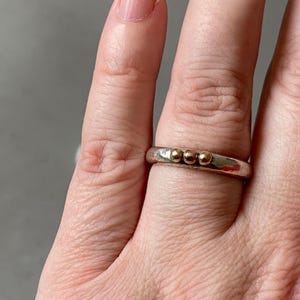 May include: A silver ring with three small gold beads. The ring is worn on a finger, showcasing the contrast between the silver band and the gold accents. The ring's design is simple and elegant.