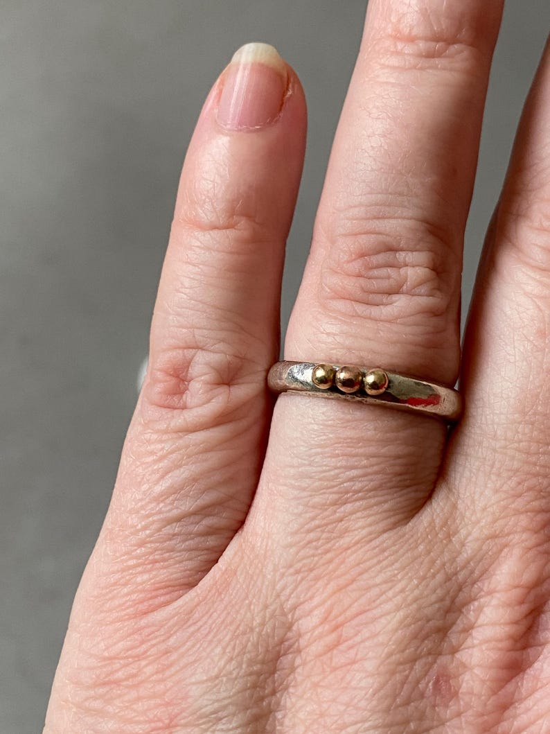 May include: A silver ring with three small gold beads. The ring is worn on a finger, showcasing the contrast between the silver band and the gold accents. The ring's design is simple and elegant.