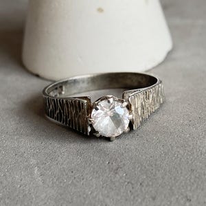 May include: A silver ring with a large, clear, round gemstone. The band has a textured, bark-like design. The ring is set against a neutral gray background with a white, cone-shaped object in the background.