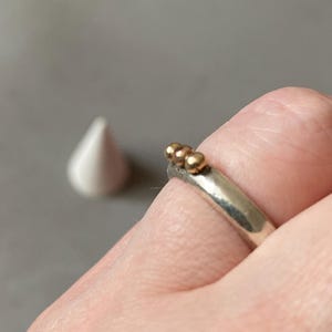 May include: A silver ring with three small gold beads on top. The ring is on a finger, with a white teardrop-shaped object in the background. The ring is a simple, elegant piece of jewelry.