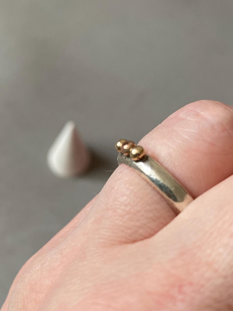 May include: A silver ring with three small gold beads on top. The ring is on a finger, with a white teardrop-shaped object in the background. The ring is a simple, elegant piece of jewelry.
