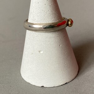 May include: A silver ring with a small gold accent, displayed on a white, cone-shaped stand. The ring has a simple, classic design with a smooth, polished band and a small, round gold detail. The stand provides a neutral backdrop, highlighting the ring's features.