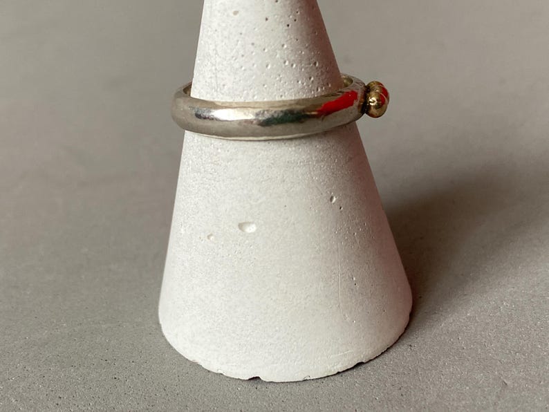 May include: A silver ring with a small gold accent, displayed on a white, cone-shaped stand. The ring has a simple, classic design with a smooth, polished band and a small, round gold detail. The stand provides a neutral backdrop, highlighting the ring's features.