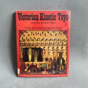 May include: A vintage book titled "Victorian Kinetic Toys and How to Make Them" by Philip and Caroline Freeman Sayer. The cover is red with black text and illustrations of antique toys. The book is a guide to creating Victorian-era kinetic toys.