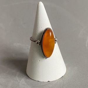 Puede incluir: Anillo de plata con una piedra ovalada de color ámbar. La piedra está engastada en un bisel detallado con pequeñas cuentas de plata a cada lado. El anillo se exhibe sobre un soporte blanco en forma de cono.