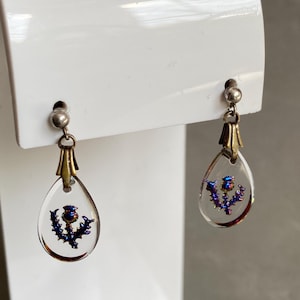 May include: A pair of drop earrings featuring clear, teardrop-shaped pendants. Each pendant has a blue and purple thistle design. The earrings have silver-colored posts and brass-colored decorative caps.