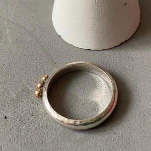 May include: A silver ring with three small gold beads clustered on one side. The ring has a hammered texture and a simple, elegant design. The ring is shown on a gray surface.