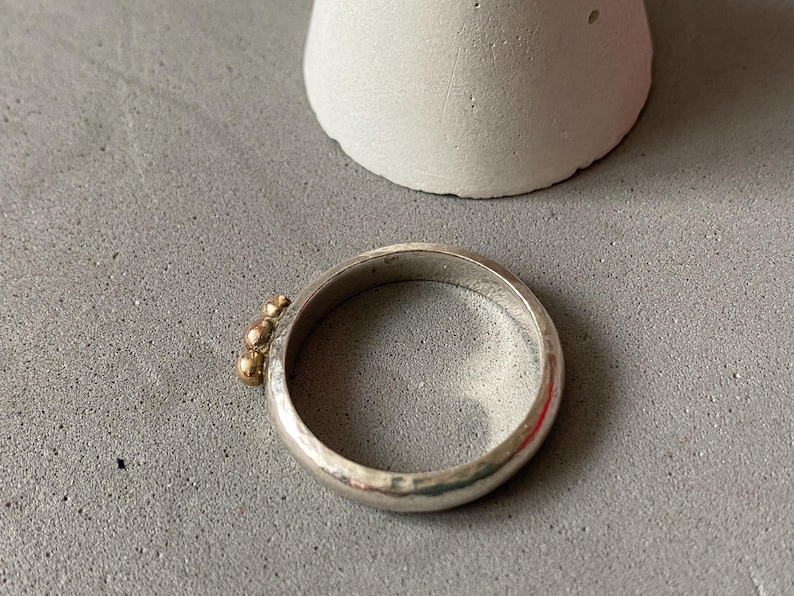 May include: A silver ring with three small gold beads clustered on one side. The ring has a hammered texture and a simple, elegant design. The ring is shown on a gray surface.