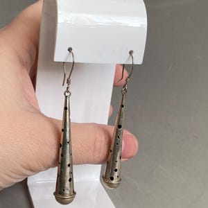 以下が含まれることがあります： シルバーカラーのぶら下がりピアス。各イヤリングは、小さな穴が均等に配置された円錐形をしています。イヤリングはシンプルなフックから吊り下げられ、白いスタンドに展示されています。