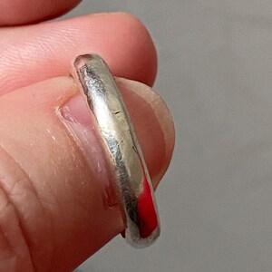 May include: A silver ring with a smooth, rounded band. The ring is held between fingers, showcasing its polished surface and slight imperfections. The ring's design is simple and classic, suitable for everyday wear.