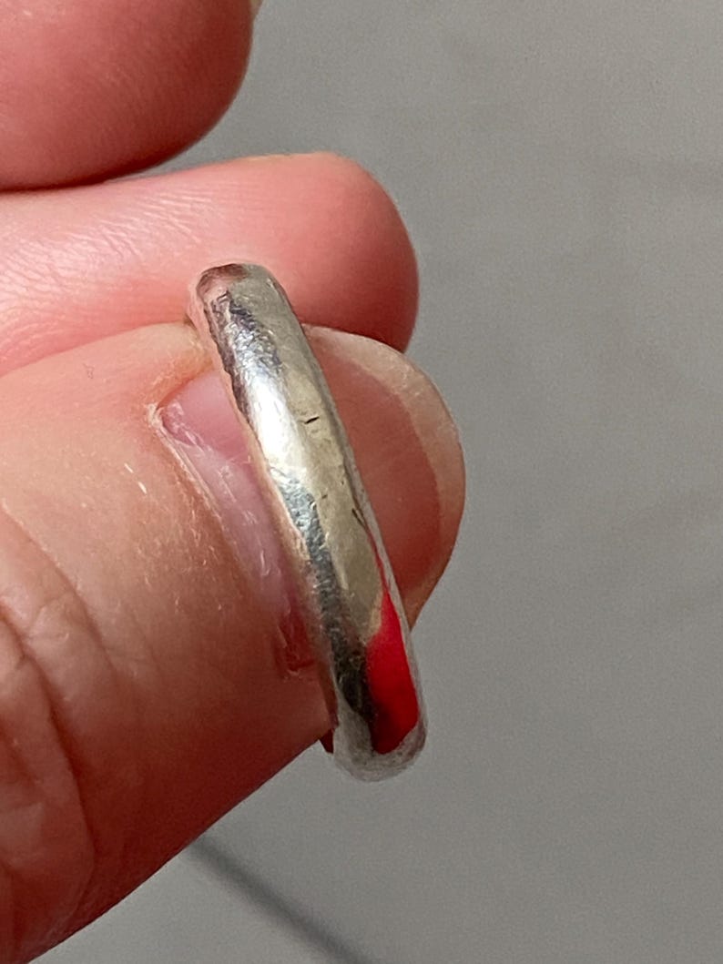 May include: A silver ring with a smooth, rounded band. The ring is held between fingers, showcasing its polished surface and slight imperfections. The ring's design is simple and classic, suitable for everyday wear.