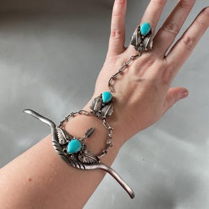 以下が含まれることがあります： ターコイズストーンとチェーンがリングにつながったシルバーブレスレット。ブレスレットは湾曲したオープンエンドの形状で、複雑なディテールが施されています。