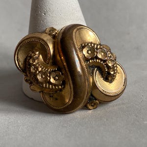 May include: A vintage gold-toned brooch with an ornate, swirling design. The brooch features floral accents and a detailed, textured surface. The piece is likely made of metal and is displayed on a white stand.
