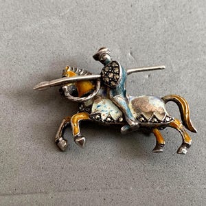 May include: A vintage brooch depicting a knight on horseback. The silver-toned brooch features a knight in blue armor with a lance, riding a horse with yellow and white enamel details. The brooch is a detailed, miniature representation.