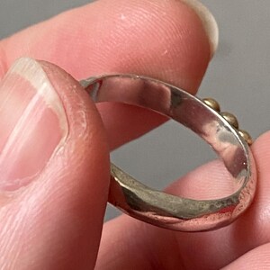 May include: A silver ring with small gold-colored beads. The ring is held between fingers, showcasing its design and texture. The band appears to be slightly hammered, adding to its unique appearance. The ring is a piece of jewelry.