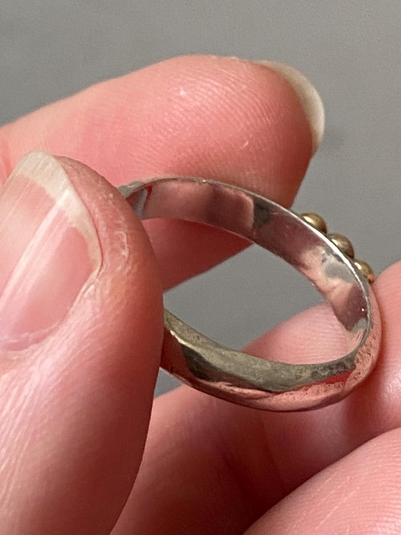 May include: A silver ring with small gold-colored beads. The ring is held between fingers, showcasing its design and texture. The band appears to be slightly hammered, adding to its unique appearance. The ring is a piece of jewelry.