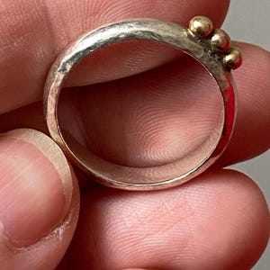 May include: A silver ring with three small gold beads clustered together. The ring has a hammered texture and a simple, elegant design. The ring is held between fingers.