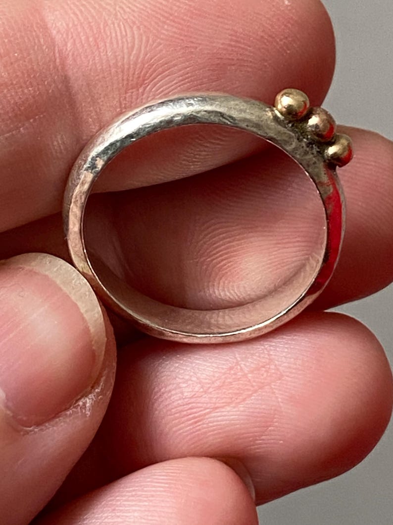 May include: A silver ring with three small gold beads clustered together. The ring has a hammered texture and a simple, elegant design. The ring is held between fingers.