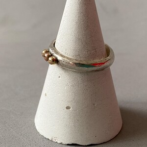 May include: A silver ring with three small gold beads clustered together. The ring is displayed on a white, cone-shaped stand. The ring has a textured, uneven surface.