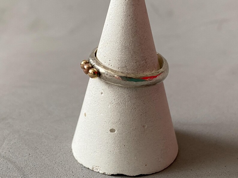 May include: A silver ring with three small gold beads clustered together. The ring is displayed on a white, cone-shaped stand. The ring has a textured, uneven surface.