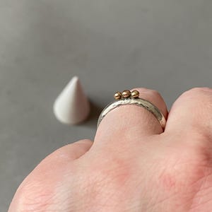 May include: A silver ring with three small gold beads. The ring has a textured band and is worn on a finger. A white, cone-shaped object is in the background against a gray backdrop.