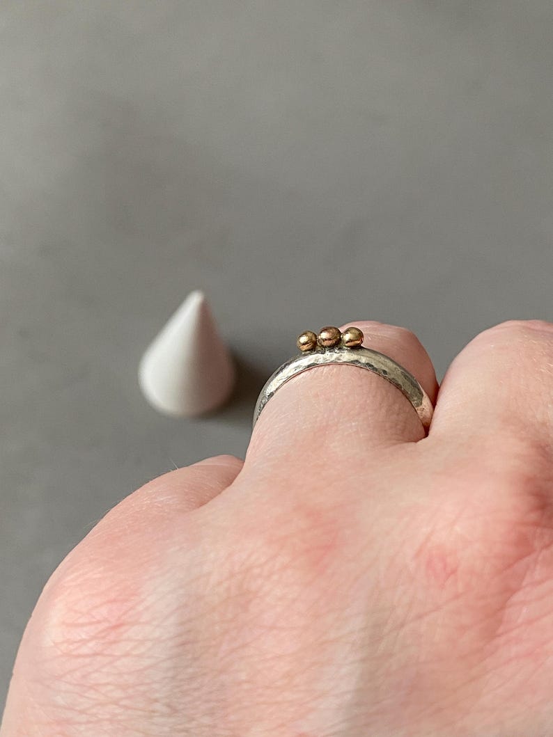 May include: A silver ring with three small gold beads. The ring has a textured band and is worn on a finger. A white, cone-shaped object is in the background against a gray backdrop.