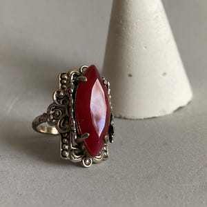 May include: A silver-toned ring featuring a large, marquise-cut deep red stone set in a decorative, filigree-style metal frame with intricate scrollwork and beaded detailing surrounding the central gemstone setting.