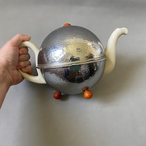 May include: A spherical teapot with a hammered silver finish. It features a white handle and spout, with small orange wooden feet and a matching knob on the lid. The teapot has a hinged lid and a silver band around the middle.