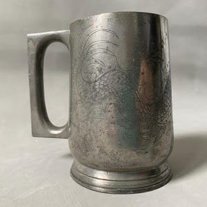 May include: A vintage silver tankard with a rectangular handle. The tankard features an etched design of a bird or floral pattern. The base of the tankard is slightly wider than the body. The tankard is made of metal and has a worn appearance.
