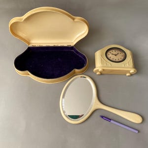 May include: A vintage vanity set featuring a cream-coloured, shell-shaped box with a purple velvet interior, a cream-coloured clock with black Roman numerals, and a cream-coloured hand mirror.